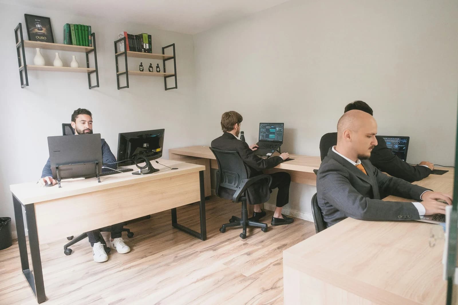 A small team of professionals working on laptops and desktop computers at wooden desks in a bright office, demonstrating how small business teams scale their capacity.