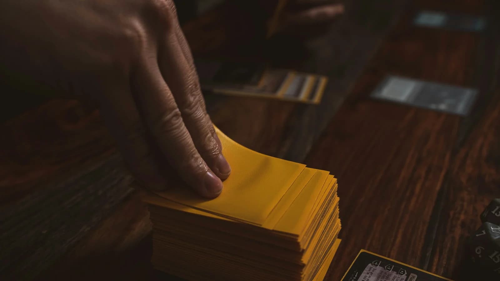 Close-up of a person's hand drawing a card from a thick stack of yellow-sleeved tabletop game cards on a wooden surface, conceptualizing tactical choices in GEO vs SEO.