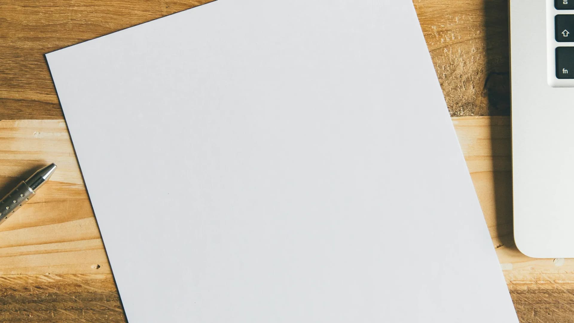 A blank sheet of paper on a wooden desk beside a pen and a laptop.