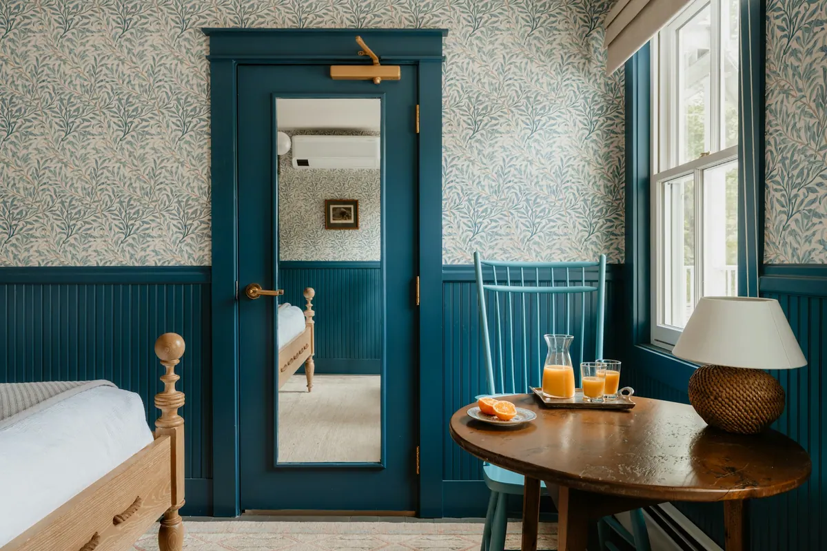 A corner guest room with patterned wallpaper, blue wainscot and a window table