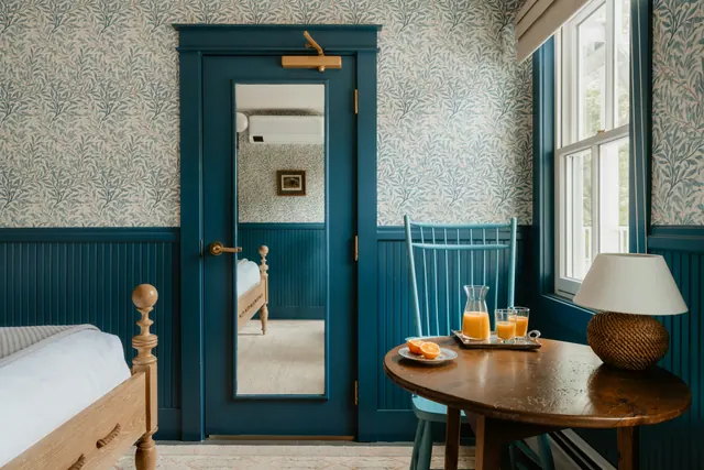 A corner guest room with patterned wallpaper, blue wainscot and a window table
